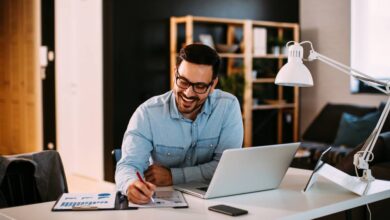 A young professional working at his home office—he productively writes on a pad of paper while on his laptop.