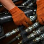 An engineer wearing orange safety gloves as they finesse a section of black hydraulic hose connections.