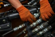 An engineer wearing orange safety gloves as they finesse a section of black hydraulic hose connections.