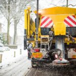 An industrial snow vehicle driving down a residential road. The vehicle is spreading street salt from behind the vehicle.