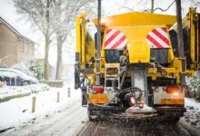 An industrial snow vehicle driving down a residential road. The vehicle is spreading street salt from behind the vehicle.