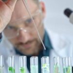 A man wearing safety goggles and a white lab coat uses a skinny glass dropper to drop a substance into several clear beakers.