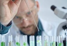 A man wearing safety goggles and a white lab coat uses a skinny glass dropper to drop a substance into several clear beakers.