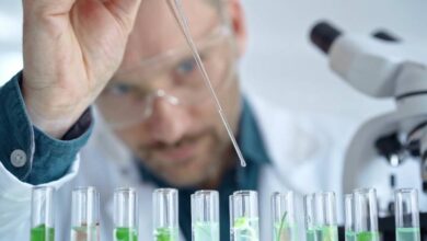 A man wearing safety goggles and a white lab coat uses a skinny glass dropper to drop a substance into several clear beakers.