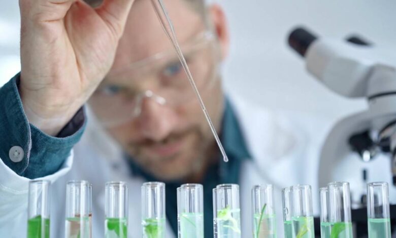 A man wearing safety goggles and a white lab coat uses a skinny glass dropper to drop a substance into several clear beakers.