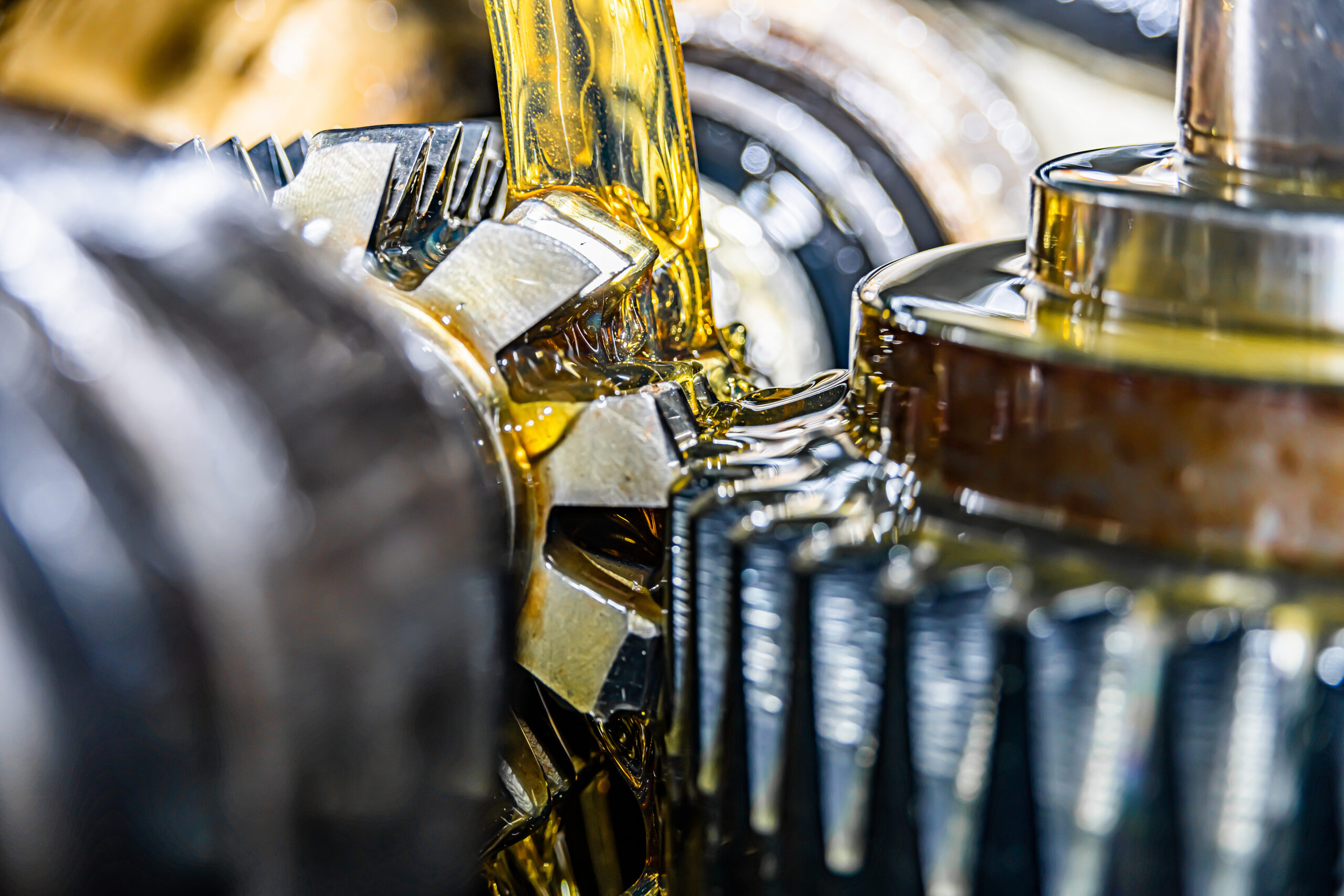 A large stream of gold oil falling onto the interlocking gears of a machine, coating their surfaces as they spin.