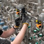 A worker wearing PPE checks a valve in a pneumatic system. There are several intricate tubes and other valves in the system.