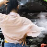 A person with one hand on their head and one on their hip looks at a car with its hood open and smoke coming out.