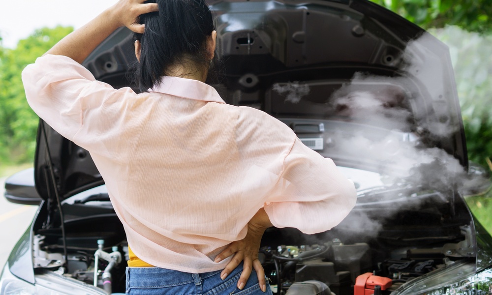 A person with one hand on their head and one on their hip looks at a car with its hood open and smoke coming out.
