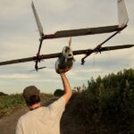 A man lifting a gray fixed-wing drone over his shoulder walks up a narrow path lined with short bushes.