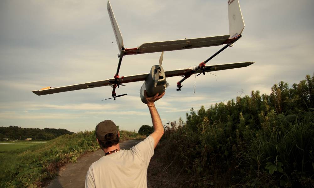 A man lifting a gray fixed-wing drone over his shoulder walks up a narrow path lined with short bushes.