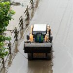 An overhead view of a skid steer operator driving the machine down a flooded roadway. Trees stand along the roadside.