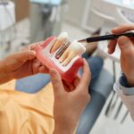 A medical model showing a dental implant. A female patient is holding the model while a doctor's hand points to it with a pen.