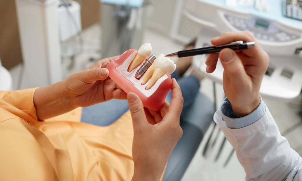 A medical model showing a dental implant. A female patient is holding the model while a doctor's hand points to it with a pen.