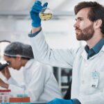 Scientist wearing a white robe and safety gloves sitting at a desk holding a glass container filled with a yellow liquid.