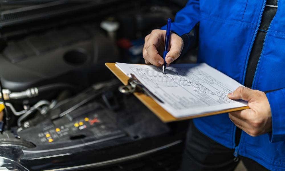 A worker in a blue jacket fills out paperwork on a clipboard with a blue pen. They are standing in front of an open car hood.