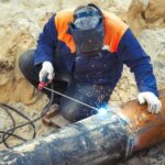 A welder fastens two underground metal pipes together using welding equipment. Hot sparks emit from their junction.