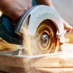 A man using a corded circular saw to rip through a piece of hardwood. The tool is spitting out sawdust.