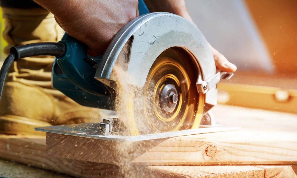 A man using a corded circular saw to rip through a piece of hardwood. The tool is spitting out sawdust.