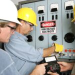 Three men wearing hard hats and safety glasses work in front of a sign that says Danger High Voltage Electricity.