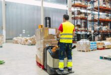 A warehouse worker driving a loaded powered pallet jack. He is wearing a yellow reflective vest and pants.