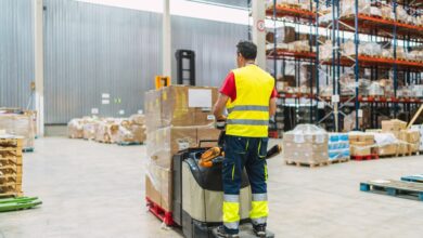 A warehouse worker driving a loaded powered pallet jack. He is wearing a yellow reflective vest and pants.