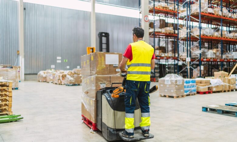A warehouse worker driving a loaded powered pallet jack. He is wearing a yellow reflective vest and pants.