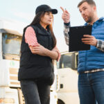 A man with a tablet points into the distance while talking to a female trucker next to him. Behind them are three semi-trucks.