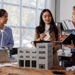 Three women inside an office stand around a large desk with a model of a building. One woman holds a model wind turbine.