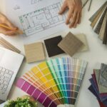 An overhead view of a desk with a fan deck, square flooring samples, a laptop, and a pencil drawing of a home's layout.
