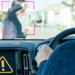 A man driving a car has an alert on his dashboard as a young woman is walking in front of his car on a sunny day.
