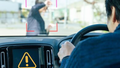 A man driving a car has an alert on his dashboard as a young woman is walking in front of his car on a sunny day.