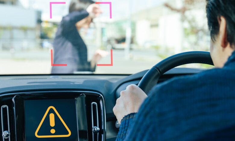 A man driving a car has an alert on his dashboard as a young woman is walking in front of his car on a sunny day.