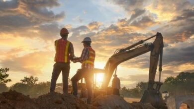 Two construction workers wearing safety vests and hard hats standing on a mound of dirt in front of an excavator.