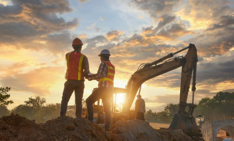 Two construction workers wearing safety vests and hard hats standing on a mound of dirt in front of an excavator.