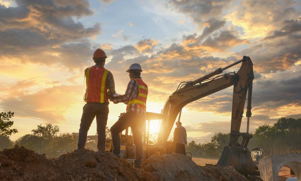 Two construction workers wearing safety vests and hard hats standing on a mound of dirt in front of an excavator.