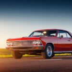 A classic red car with two doors sits on a two-lane road next to an open field. The sunshine is hitting the car.