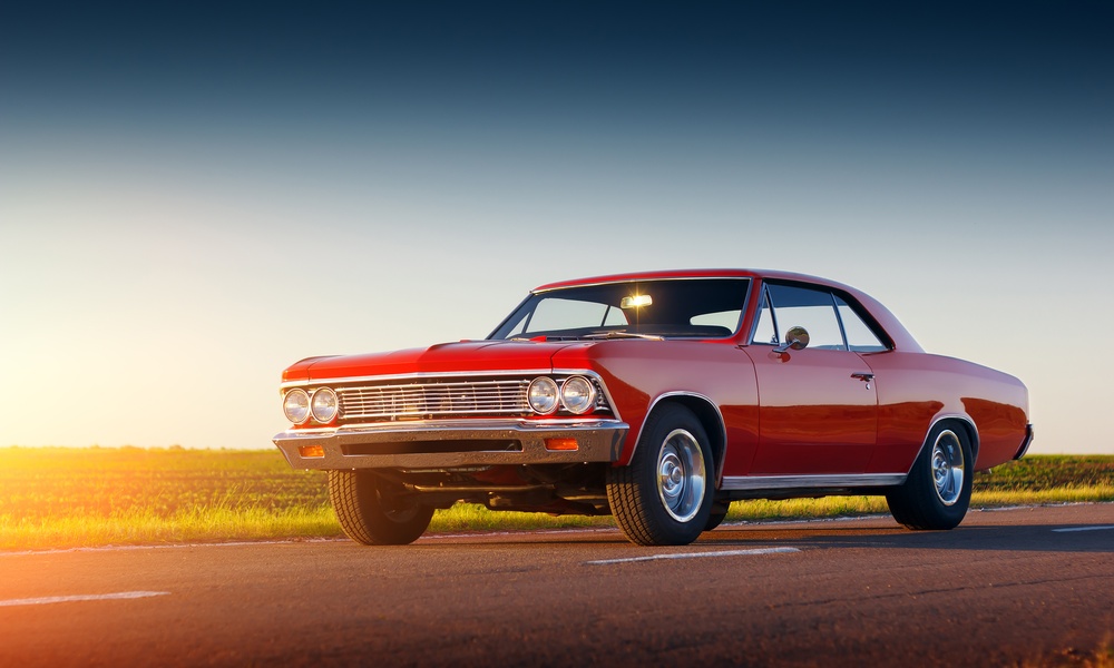 A classic red car with two doors sits on a two-lane road next to an open field. The sunshine is hitting the car.
