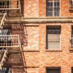 The exterior of a red brick building with a light-colored fire escape. Some of the windows are covered with bars.