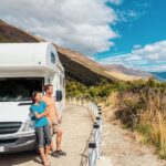A young man and woman lean back against the hood of a parked RV and look at a grassy coast. Mountains are behind them.