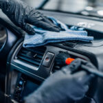 A close-up of a person’s hands wearing black gloves using a brush a cloth to clean a black dashboard.