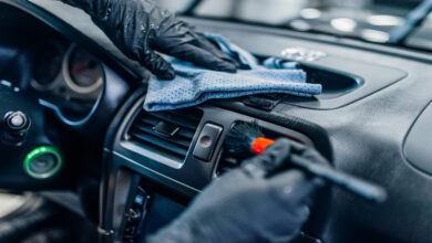 A close-up of a person’s hands wearing black gloves using a brush a cloth to clean a black dashboard.