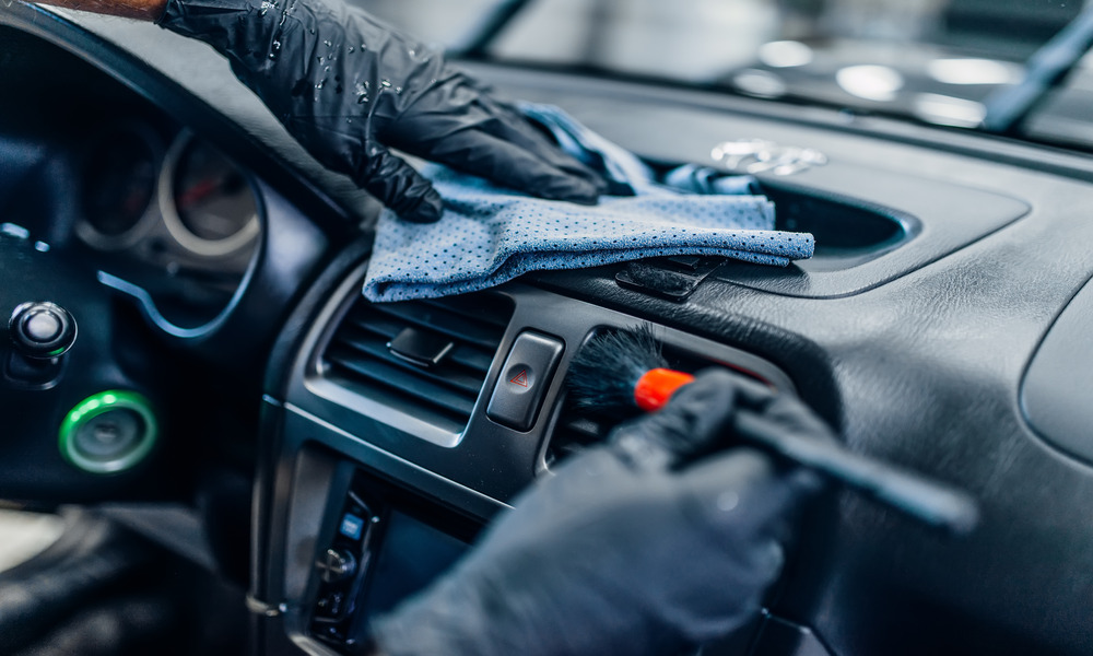 A close-up of a person’s hands wearing black gloves using a brush a cloth to clean a black dashboard.