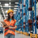 A smiling woman in an orange safety vest, jeans, and a yellow hard hat stands in a warehouse holding a clipboard.