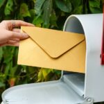 A person's hand retrieving a manila envelope from a white mailbox with a red flag surrounded by greenery.