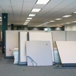 An office with multiple gray cubicles on gray carpeting. Stacks of drywall sheets rest on the cubicle walls.