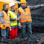 Three workers wearing hard hats and high-visibility vests stand together at a mining quarry and look off to the side.