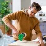 A young man wearing a hoodie uses a tape gun to secure a cardboard box in his living room.