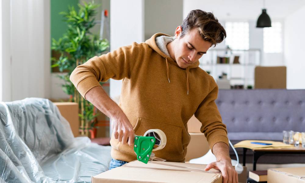 A young man wearing a hoodie uses a tape gun to secure a cardboard box in his living room.