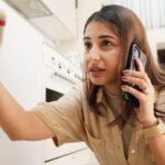 A concerned woman holds a white and red towel to stop a leak under her sink while on a phone call.